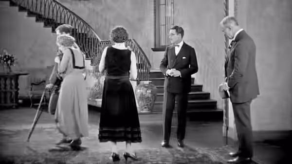 A woman with a cane stands with three men in a grand staircase hall