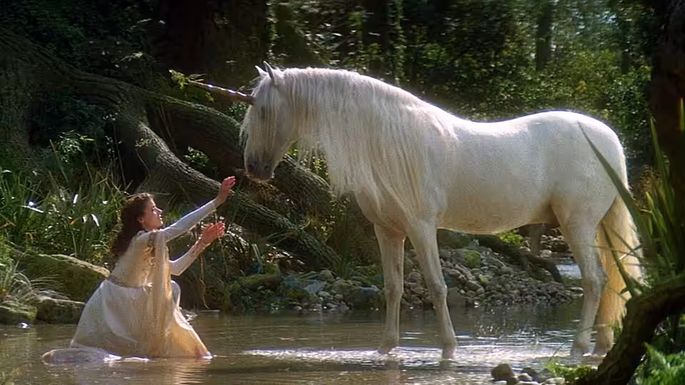 Princess Lili (Mia Sara) reaches toward a white unicorn standing in a forest stream