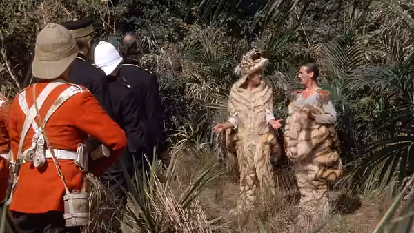 Men in dinosaur costumes stand before soldiers in a tropical jungle environment