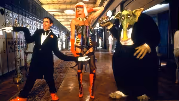 A man in a tuxedo poses with a pink-haired woman and a green alien creature in a locker room