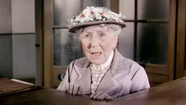 Mrs. Wilberforce (Katie Johnson) sits behind a desk, wearing a flower-trimmed hat
