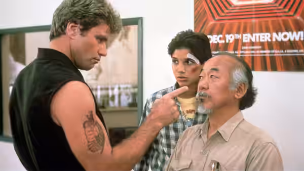 Miyagi (Pat Morita) being pointed at by a man as a woman watches behind