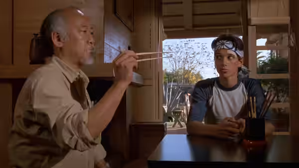 Miyagi (Pat Morita) eating with chopsticks while Daniel (Ralph Macchio) watches across the table