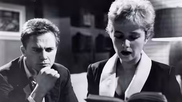 Man and woman sit closely reading from open book in dark interior setting