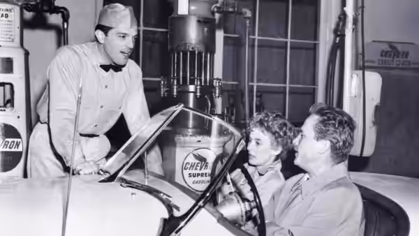Attendant in cap leans over race car talking to couple inside at gas station