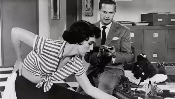 Woman in striped top leans over desk reaching forward while man examines small object