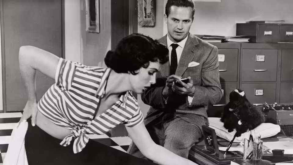 Woman in striped top leans over desk reaching forward while man examines small object