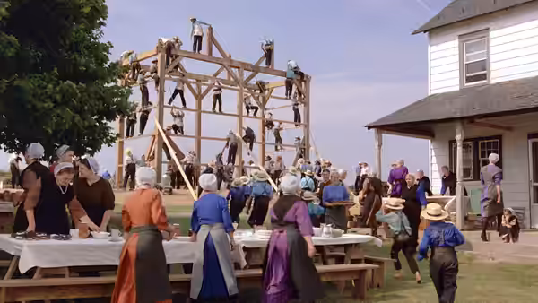 Amish women prepare food at long tables while men raise a wooden barn frame