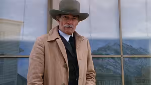 Stockburn (John Russell) standing before a glass building with mountain reflection