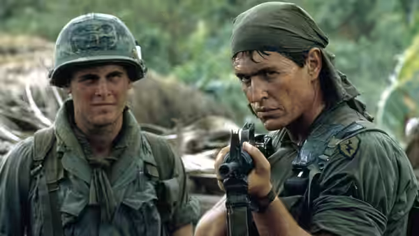 Sergeant Barnes checks his weapon beside a helmeted soldier in camp