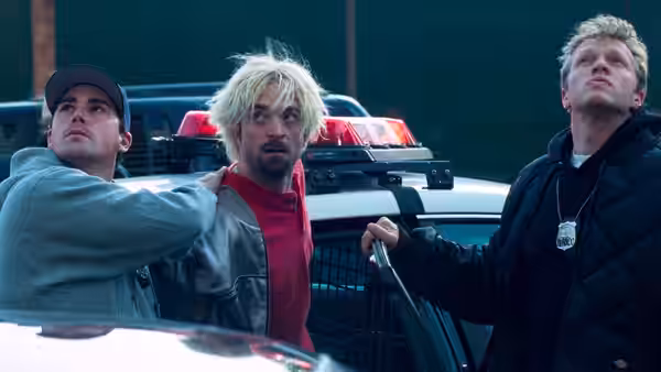 Nick Nikas in a red jacket stands between two men beside a police car