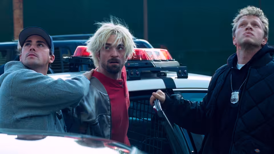 Nick Nikas in a red jacket stands between two men beside a police car