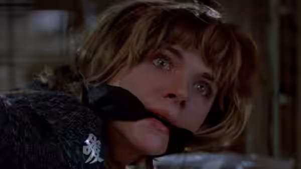 A blonde woman with a gag looks upward with wide eyes