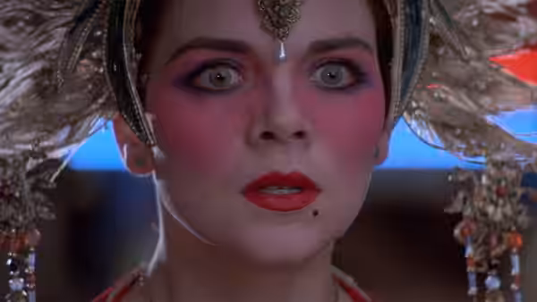 A woman with an ornate headdress stares forward with vivid makeup