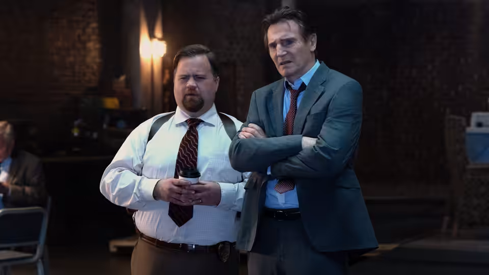 Frank Drebin Jr. (Liam Neeson) stands with arms crossed beside Ed Hocken Jr. (Paul Walter Hauser)