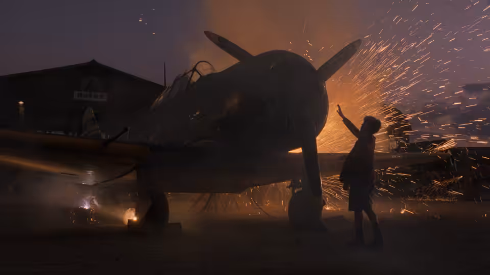 boy in school cap stands near a burning plane as sparks fly at night