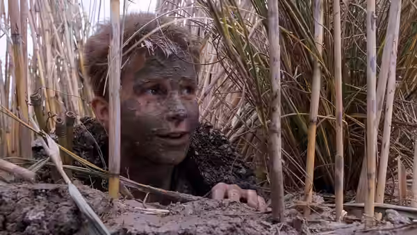 Jim (Christian Bale) lies muddy among reeds with face and hair caked in dirt