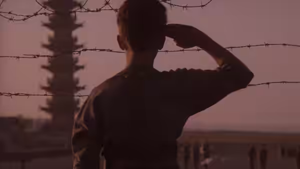 boy in uniform salutes with his back to camera before barbed wire and a pagoda
