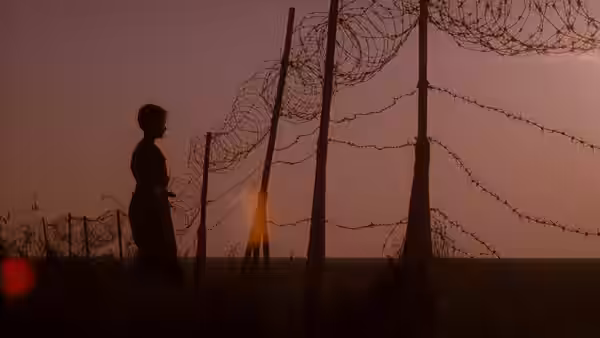 silhouetted figure walks past tall barbed wire fences against a sunset sky