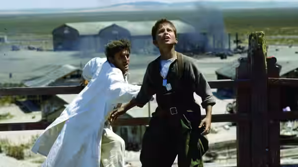 two men grapple beside a wooden fence in a dusty airfield with hangars