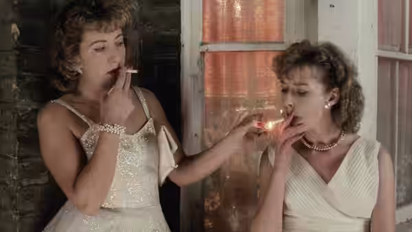Two women in evening dresses light cigarettes by a window