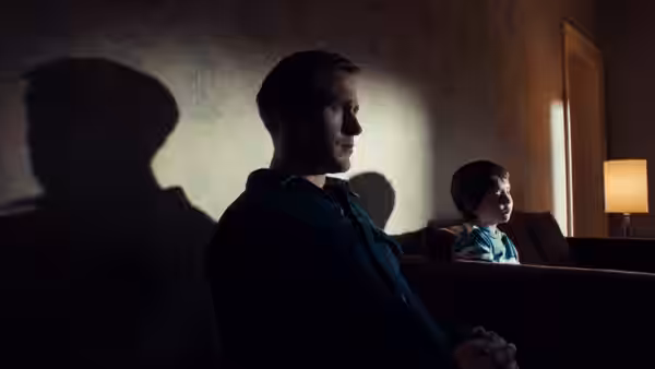 Driver (Ryan Gosling) sits on a couch beside a young boy in a shadowed room