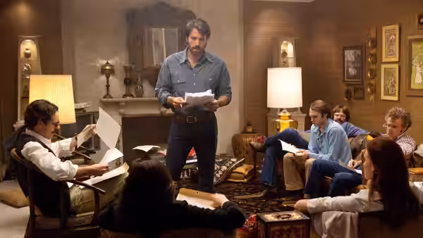 Tony Mendez stands holding papers while colleagues review documents in a living room