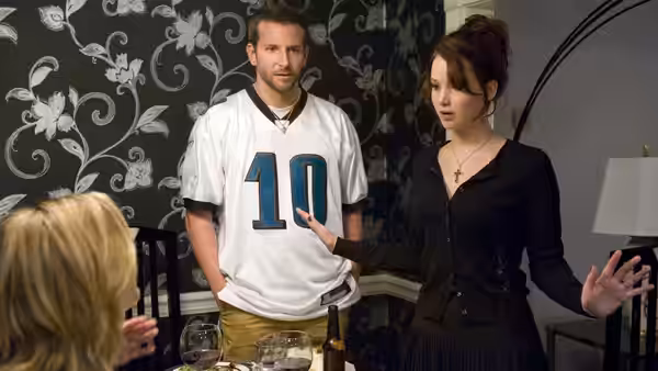 Pat (Bradley Cooper) watching Tiffany (Jennifer Lawrence) gesture in a dining room