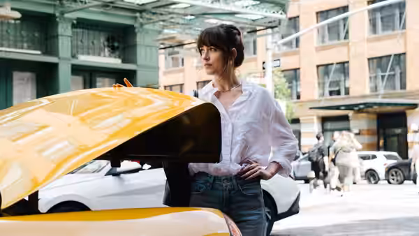 Lucy (Dakota Johnson) standing beside a yellow taxi with its hood open