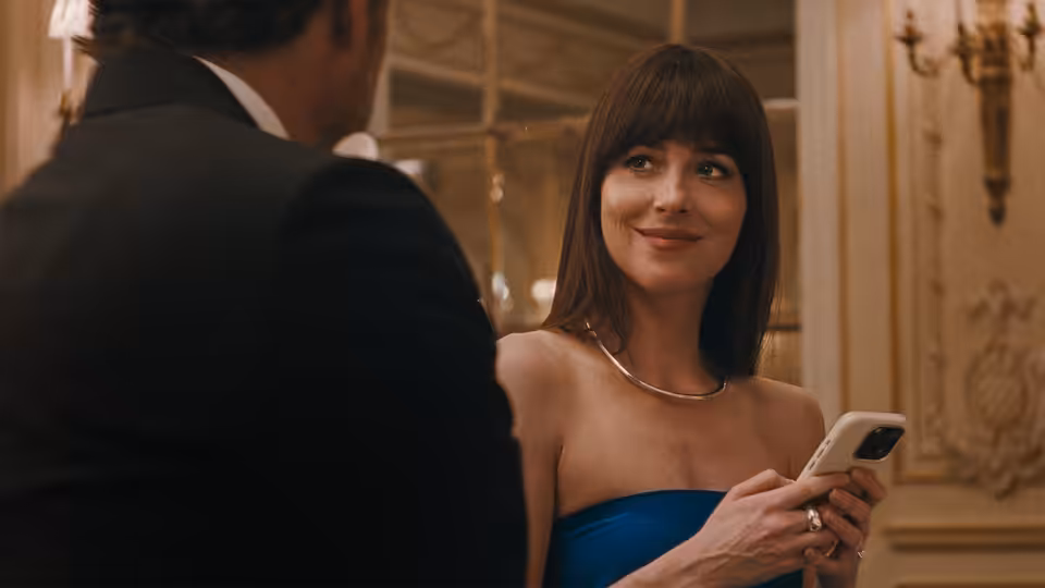 Lucy (Dakota Johnson) holding a phone while speaking to a man in a tuxedo