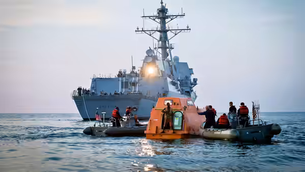 A naval warship approaches an orange lifeboat on the open sea