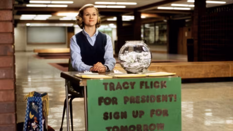 Tracy Flick (Reese Witherspoon) stands behind her campaign table in a school hallway
