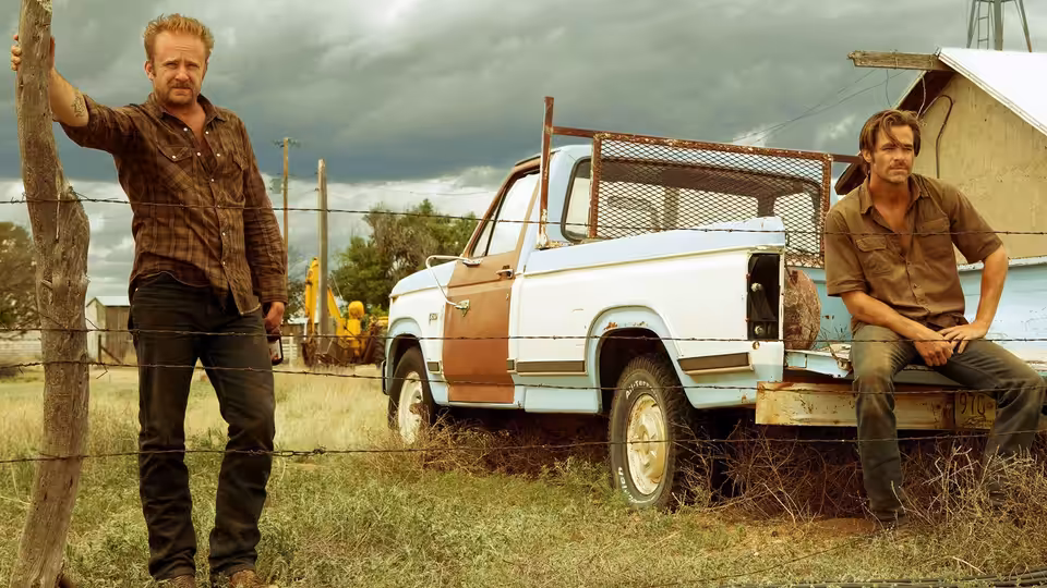 Jeff Bridges stands by a fence next to a truck with Chris Pine seated on it