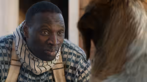 Perrault (Omar Sy) leans toward a dog outside a wooden building