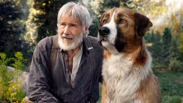 John Thornton (Harrison Ford) sits beside Buck in a forest clearing