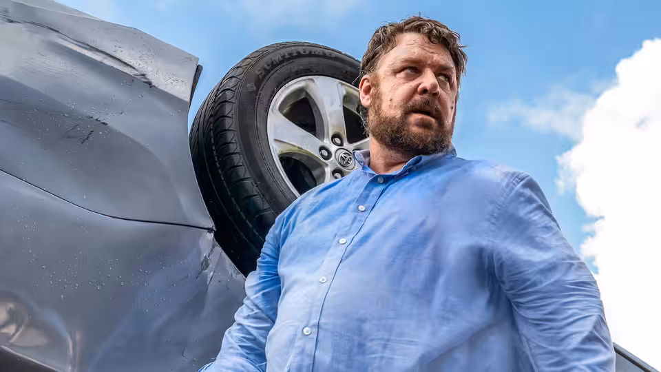 Tom Cooper (Russell Crowe) stands beside an overturned car, looking over his shoulder