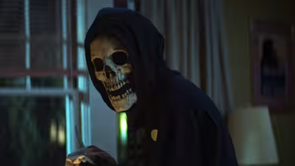 A skull-masked figure in a dark hood stands inside a room