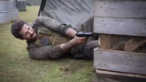 Jake Harris (Scott Adkins) lies on the grass and aims a pistol around wooden cover