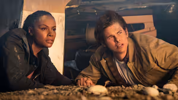 Rachel and Tom crawl beneath a vehicle, peering out from under its chassis