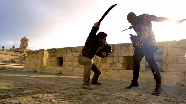Mike Fallon (Scott Adkins) raises a machete against an armed man on a stone rooftop