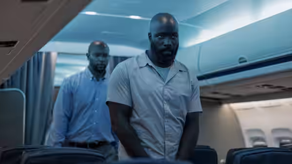 Louis Gaspare (Mike Colter) stands handcuffed in an airplane cabin beside another prisoner