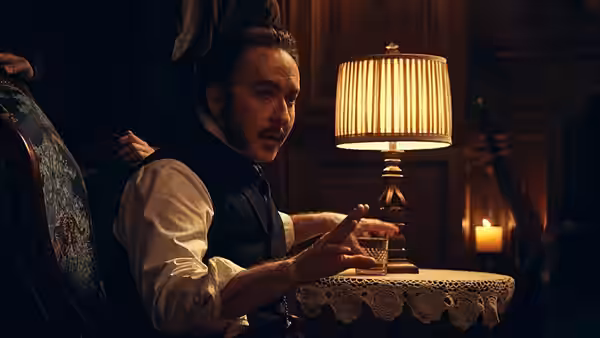 A mustached man gestures from his chair beside a lit table lamp