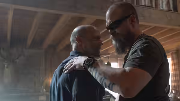 Levon Cade (Jason Statham) stands close to a bearded man in a rustic room