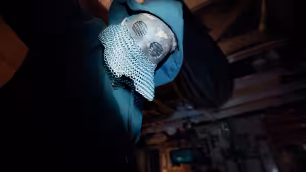 A hooded figure wearing a metal face covering leans beneath wooden rafters