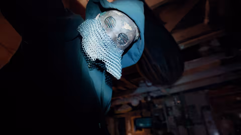 A hooded figure wearing a metal face covering leans beneath wooden rafters