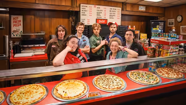 A group of pizza shop workers pose behind the counter beside pizzas on display