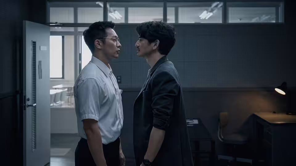 A man in a white shirt faces a dark-clad man in a dim office