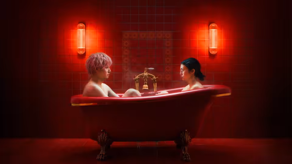 A man and woman sit facing each other in a clawfoot bathtub against red tiled walls