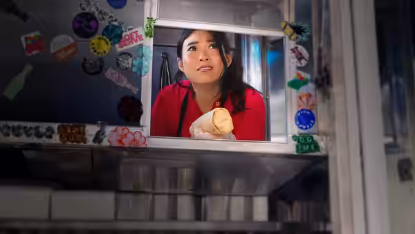 Awkwafina peers through a decorated service window, holding a wrapped sandwich