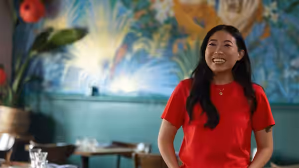 Awkwafina smiles in a red shirt inside a restaurant